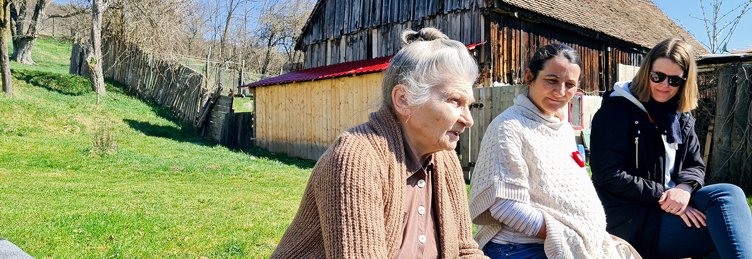 Drei Frauen unterschiedlichen Alters sitzen vor einem Holzhaus