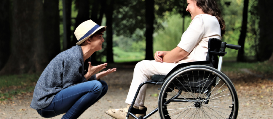 Zwei Frauen lachen sich an. Eine sitzt in der Hocke, die andere im Rollstuhl. Sie befinden sich auf einem Waldweg.