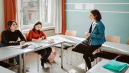 Lehrerin sitzt im Unterricht auf einem Tisch gegenüber zwei Schülerinnen